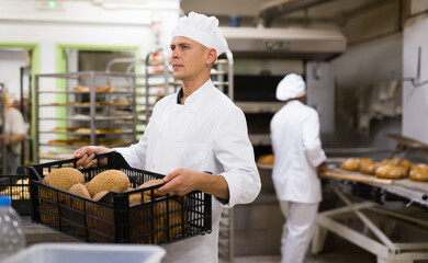 Fototapeta na wymiar Positive man worker of bakery carrying box of bread standing at kitchen