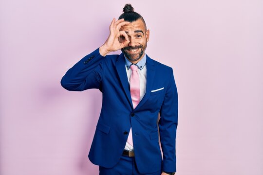 Young hispanic man wearing business suit and tie doing ok gesture with hand smiling, eye looking through fingers with happy face.