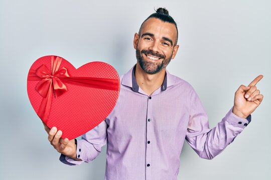 Young hispanic man holding valentine gift smiling happy pointing with hand and finger to the side