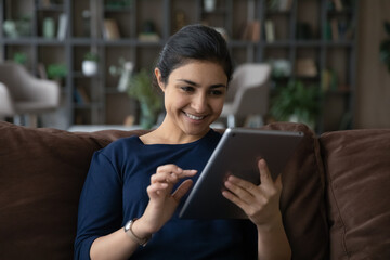 Happy young Indian ethnicity woman using digital computer tablet, enjoying web surfing information, choosing goods shopping in internet store, typing message communicating distantly online at home.