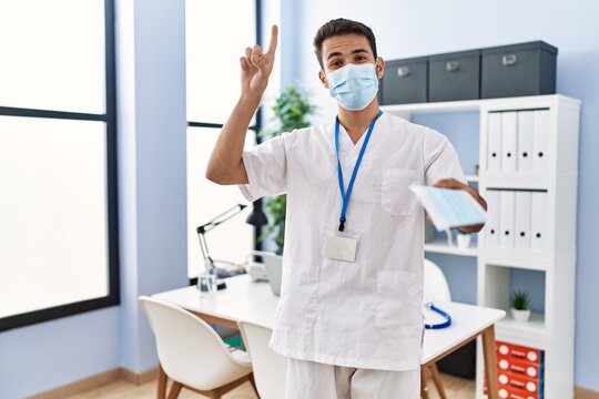 Young hispanic doctor man offering safety mask smiling with an idea or question pointing finger with happy face, number one