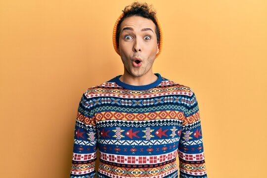 Young handsome man wearing wool hat and colorful sweater scared and amazed with open mouth for surprise, disbelief face