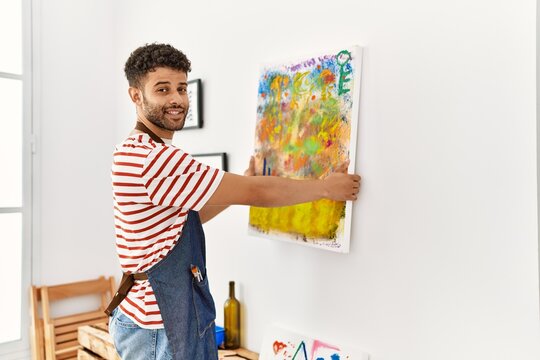 Young arab artist man smiling happy hanging draw canvas on wall at art studio.