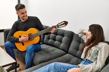 Young hispanic couple playing spanish guitar sitting on the sofa at home.