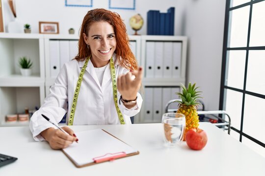 Young redhead woman nutritionist doctor at the clinic beckoning come here gesture with hand inviting welcoming happy and smiling