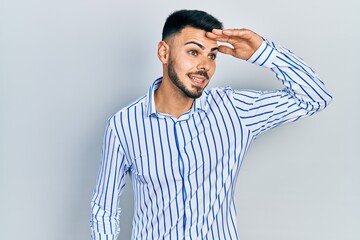 Young hispanic man with beard wearing casual striped shirt very happy and smiling looking far away with hand over head. searching concept.