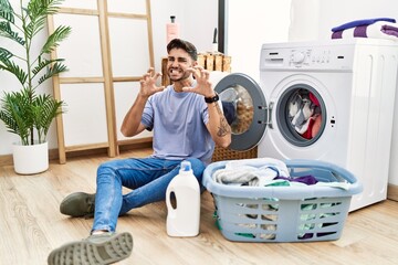 Young hispanic man putting dirty laundry into washing machine smiling funny doing claw gesture as cat, aggressive and sexy expression
