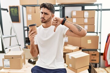 Young hispanic man working at small business ecommerce drinking a coffee with angry face, negative...