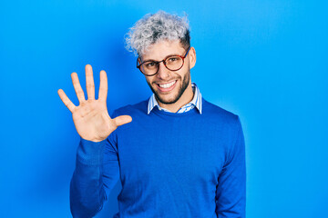 Young hispanic man with modern dyed hair wearing sweater and glasses showing and pointing up with fingers number five while smiling confident and happy.