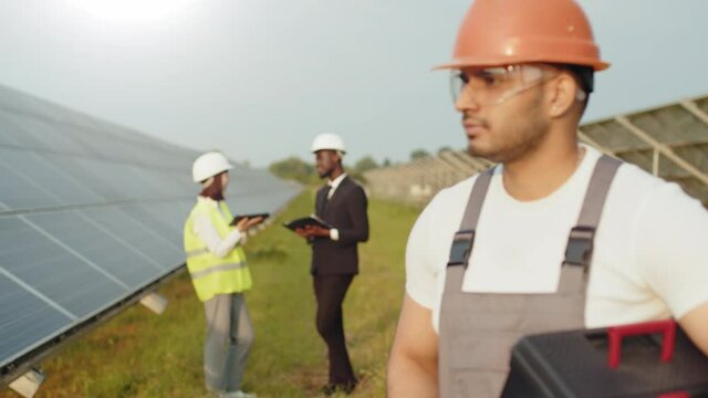 Indian man in overalls, glasses and orange helmet holding instruments and looking aside while standing among solar station. Group of engineers having discuss on background. Technician at solar farm.
