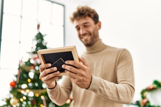 Young hispanic man holding photo standing by christmas tree at home