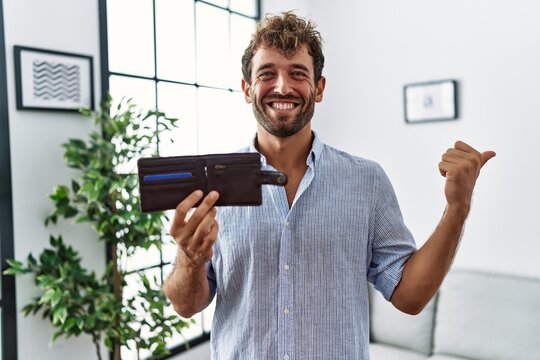 Young handsome man holding leather wallet pointing thumb up to the side smiling happy with open mouth