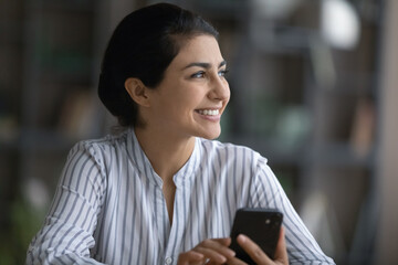 Happy young Indian businesswoman holding cellphone in hands looking in distance, feeling dreamy...