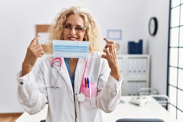 Middle age blonde woman wearing doctor uniform and medical mask winking looking at the camera with sexy expression, cheerful and happy face.