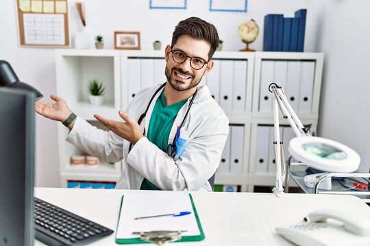 Young man with beard wearing doctor uniform and stethoscope at the clinic inviting to enter smiling natural with open hand