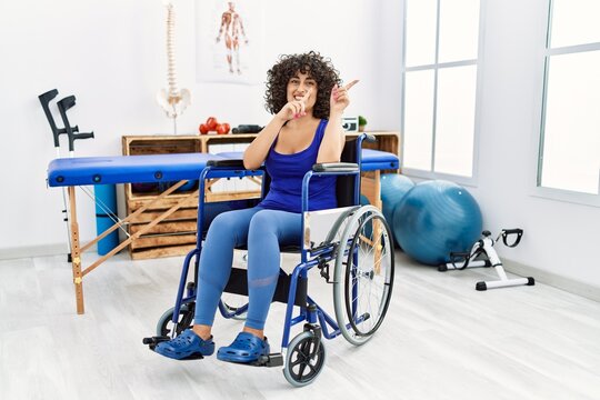 Young middle eastern woman sitting on wheelchair at physiotherapy clinic smiling and looking at the camera pointing with two hands and fingers to the side.
