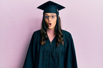 Young hispanic woman wearing graduation cap and ceremony robe afraid and shocked with surprise expression, fear and excited face.