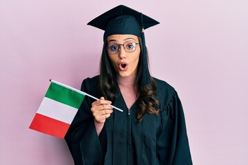 Young hispanic woman wearing graduation uniform holding italy flag scared and amazed with open mouth for surprise, disbelief face