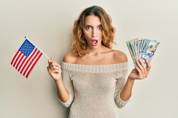 Young caucasian woman holding united states flag and dollars in shock face, looking skeptical and sarcastic, surprised with open mouth