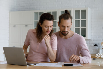 Concentrated millennial married family couple calculating domestic expenditures, analyzing paper bills, making mortgage payment in laptop e-banking application or paying for services online at home.