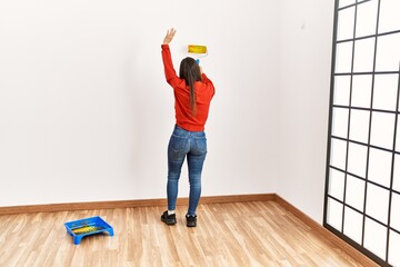 Young latin woman on back view painting wall using roller at empty room