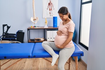Young latin woman pregnant patient sitting on massage table at clinic