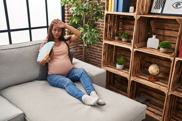 Fototapeta Young latin woman pregnant using hand fan sitting on sofa at home obraz
