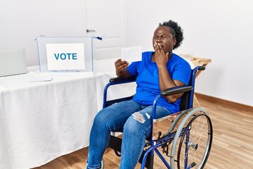 Young african woman sitting on wheelchair voting putting envelop in ballot box bored yawning tired...