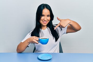 Beautiful hispanic woman with nose piercing drinking a cup coffee smiling happy pointing with hand...