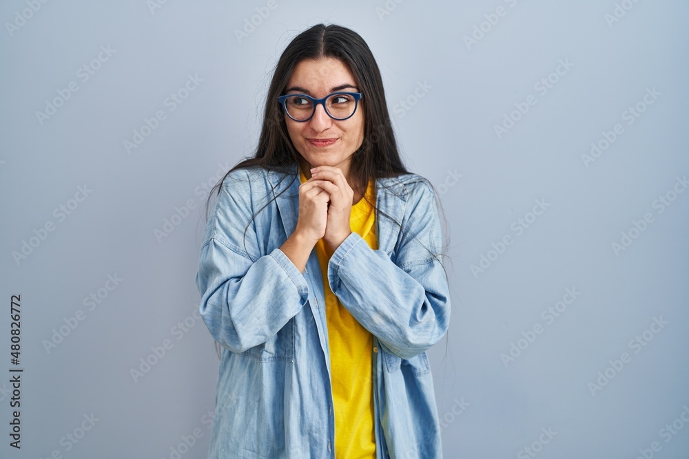 Poster Young hispanic woman standing over blue background laughing nervous and excited with hands on chin looking to the side