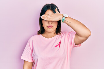 Fototapeta na wymiar Young hispanic woman wearing pink cancer ribbon on t shirt covering eyes with hand, looking serious and sad. sightless, hiding and rejection concept