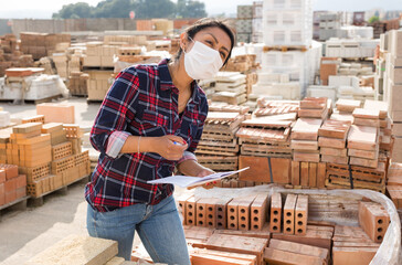 Female worker in protective face mask checking quantity of red bricks in warehouse of building materials
