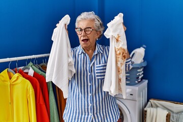 Senior woman with grey hair holding clean white t shirt and t shirt with dirty stain winking...