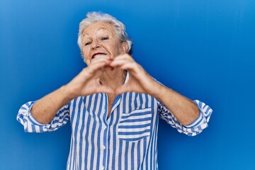 Senior woman with grey hair standing over blue background smiling in love doing heart symbol shape with hands. romantic concept.