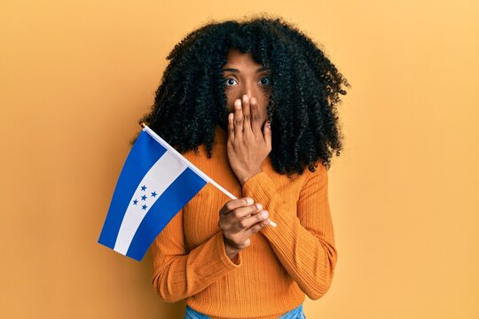 African american woman with afro hair holding honduras flag covering mouth with hand, shocked and afraid for mistake. surprised expression