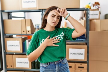 Young brunette woman wearing volunteer t shirt at donations stand touching forehead for illness and...