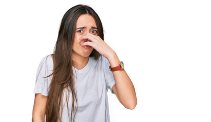 Young hispanic girl wearing casual white t shirt smelling something stinky and disgusting, intolerable smell, holding breath with fingers on nose. bad smell