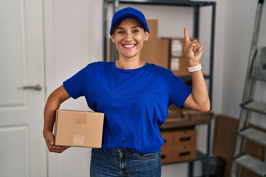 Middle age brunette woman working wearing delivery uniform and cap smiling amazed and surprised and pointing up with fingers and raised arms.