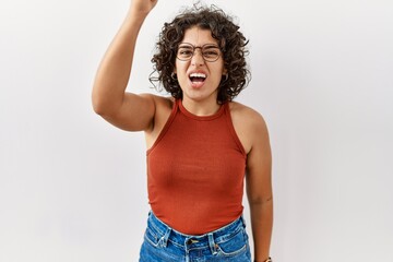Young hispanic woman wearing glasses standing over isolated background angry and mad raising fist frustrated and furious while shouting with anger. rage and aggressive concept.