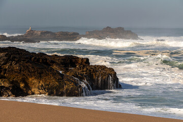 Landscape of the picturesque sandy coast of the Atlantic Ocean with foaming waves and large brown stones, an old lighthouse, blue water and a clear sky, Portugal, Porto