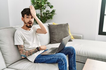 Hispanic man with beard sitting on the sofa surprised with hand on head for mistake, remember...