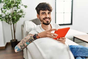Fototapeta premium Young hispanic man using touchpad sitting on the sofa at home
