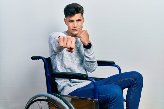 Young hispanic man sitting on wheelchair punching fist to fight, aggressive and angry attack, threat and violence