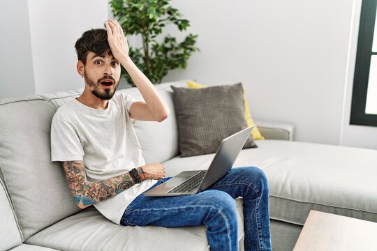 Hispanic man with beard sitting on the sofa surprised with hand on head for mistake, remember error. forgot, bad memory concept.