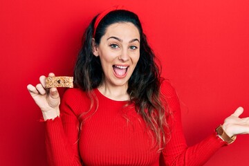 Fototapeta premium Young hispanic woman eating protein bar as healthy energy snack celebrating achievement with happy smile and winner expression with raised hand