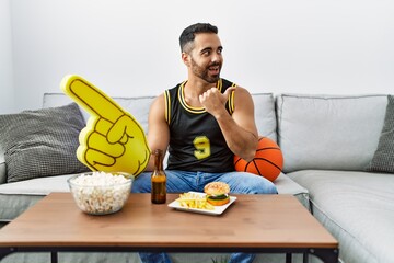 Young hispanic man with beard holding basketball ball cheering tv game pointing thumb up to the side smiling happy with open mouth