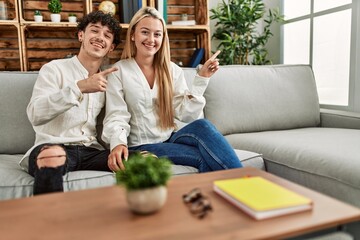 Young beautiful couple sitting on the sofa at home smiling happy pointing with hand and finger to the side