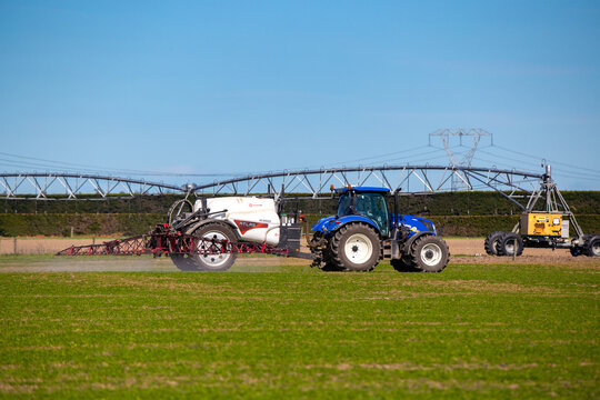 Darfield, Canterbury, New Zealand, September 11 2021: A farmer with a blue New Holland tractor, sprays one of his fields in spring. There is also a central pivot irrigator on the farm.