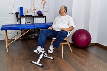 Senior caucasian man at physiotherapy clinic using pedal exerciser looking to side, relax profile pose with natural face with confident smile.