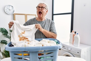 Senior caucasian man holding dirty t shirt with stain angry and mad screaming frustrated and furious, shouting with anger looking up.